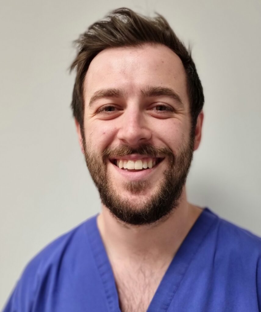 An image of Dan Nicholls, a white man with short dark hair and beard, wearing blue scrubs and smiling at the camera.