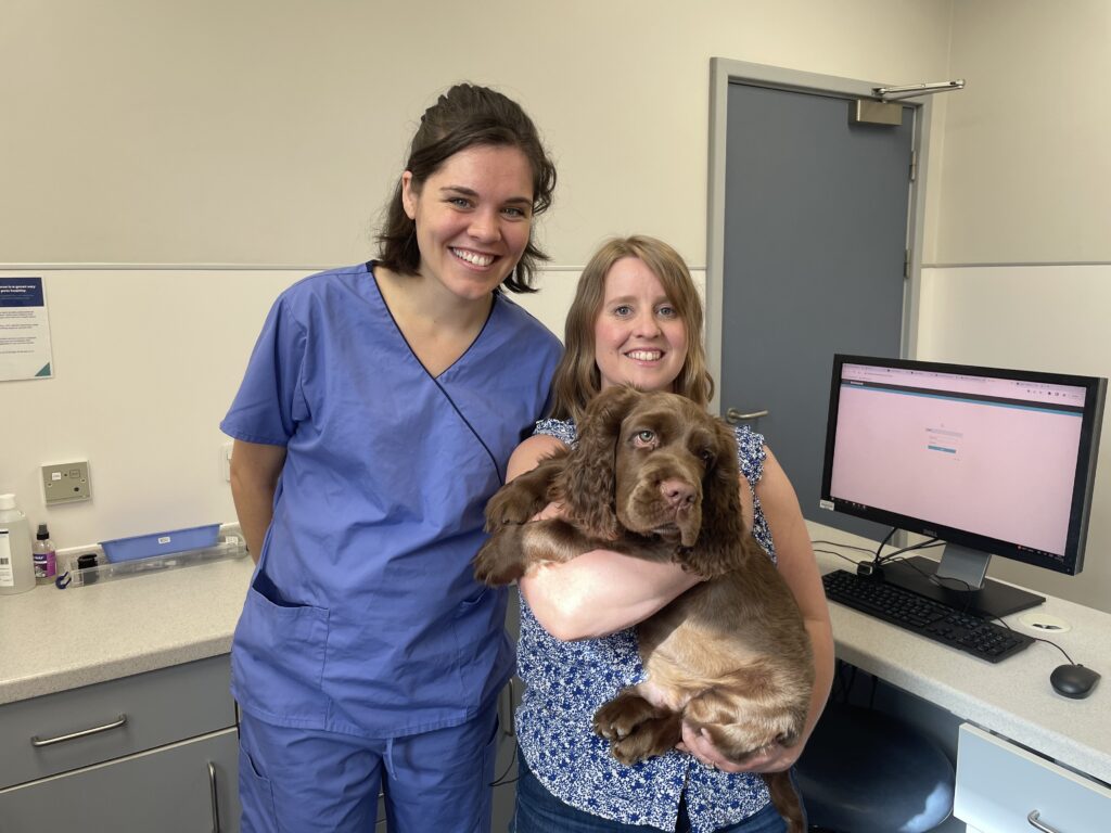 An image of Elton, a brown Sussex spaniel, being held by his owner, Philippa, a white woman with brown hair. Standing next to Philippa is Holly, a white woman with dark hair wearing blue scrubs.