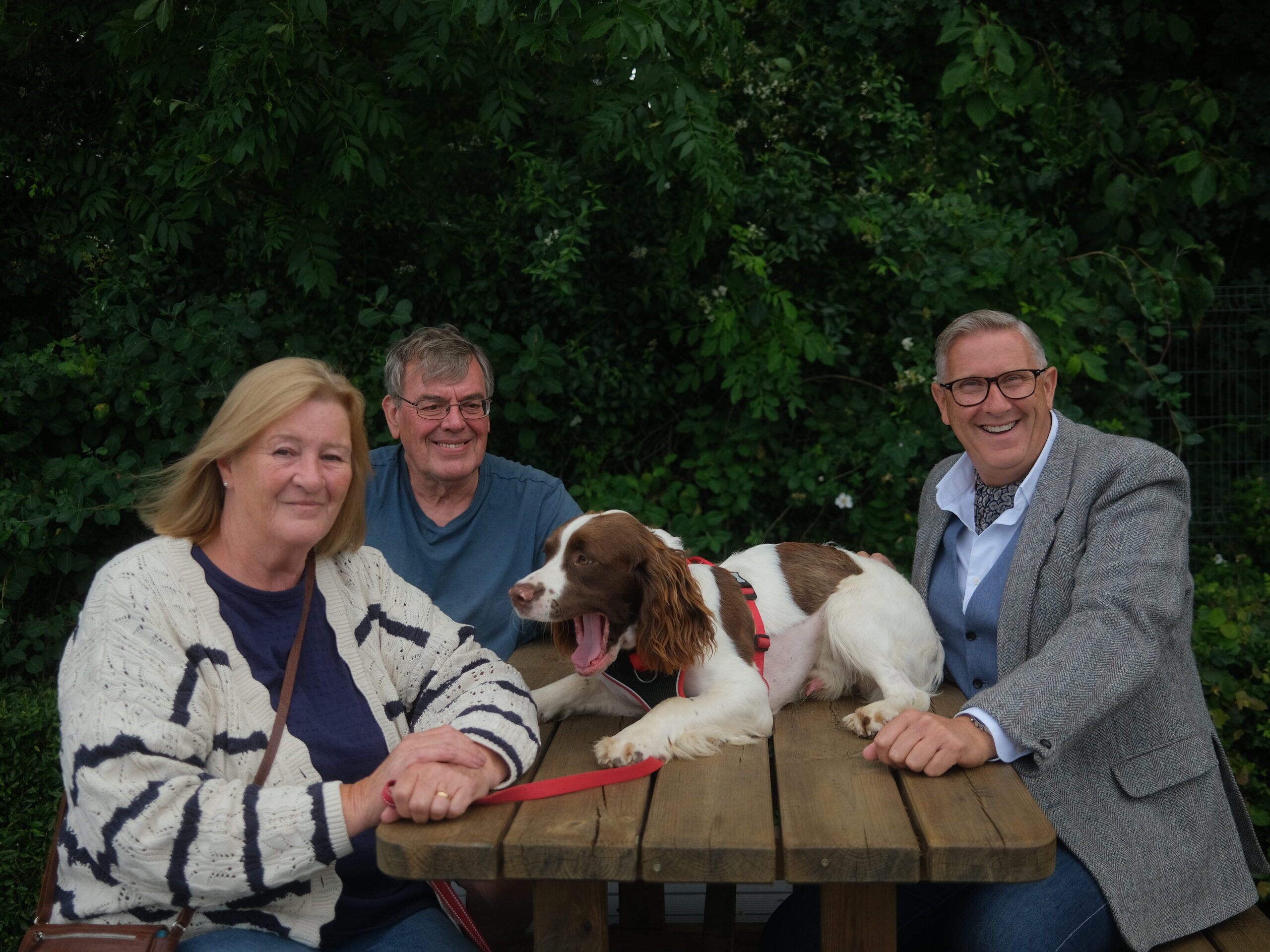 An image of Louie, a springer spaniel dog, sat on a picnic table. Seated to the left is Louie's owners, Kevin and Nicky Blythe, to the right is Graeme Hall, presenter of the Dog Hospital with Graeme Hall