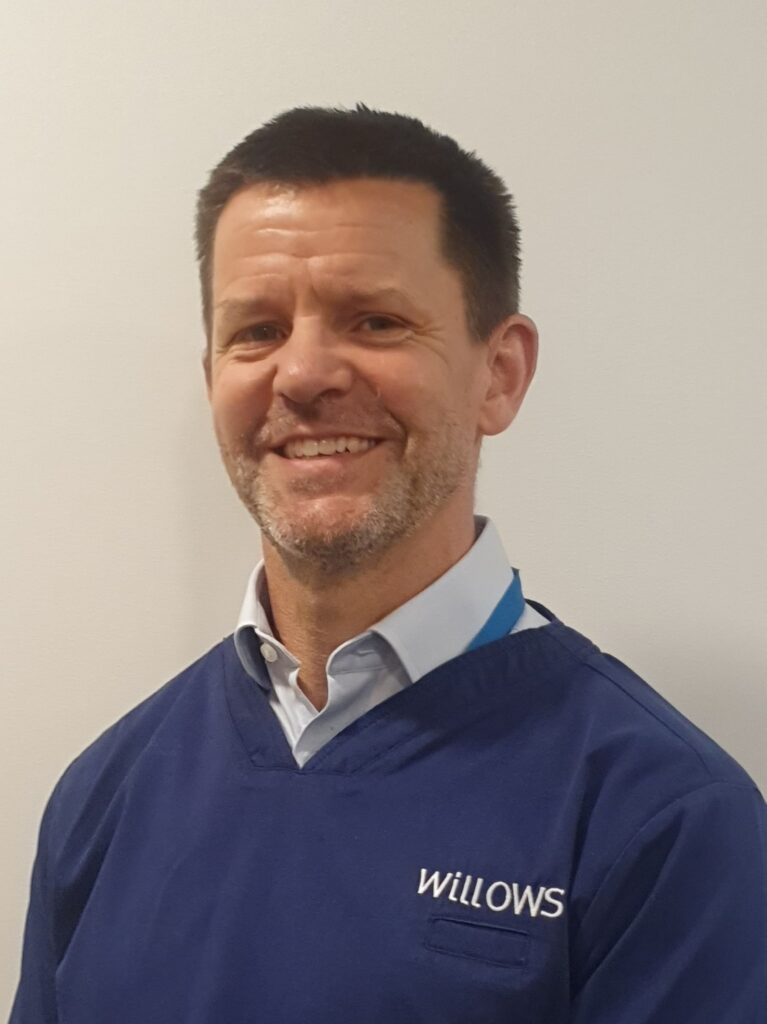 An image of Orthopaedic specialist Toby Gemmill, a white man with dark hair. He is wearing a dark blue scrub top with a light blue shirt underneath. He is looking at the camera and smiling.