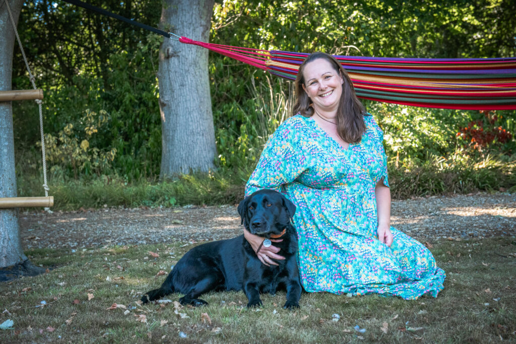 An image of a black Labrador lying on the grass in a garden, kneeling next to him is his owner, a white woman with brown hair who is wearing a dress with a floral pattern. She is smiling and has one arm around the Labrador