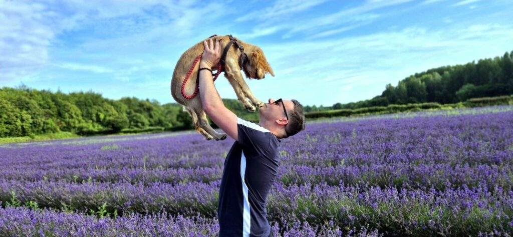 An image of a white man wearing a t-shirt and sunglasses standing in a large lavender field holding up a puppy.