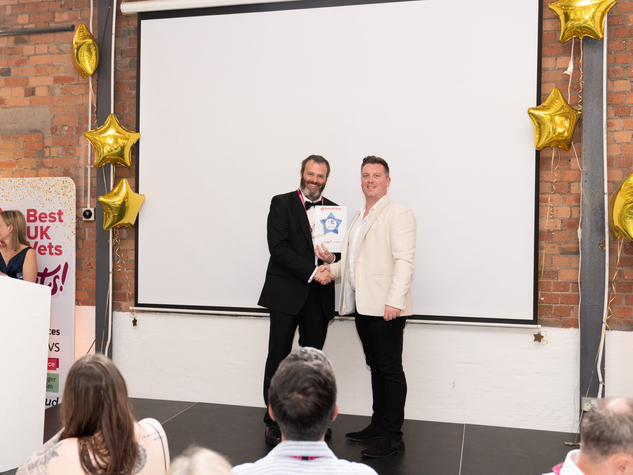 An image of two white men standing on a stage which is decorated with gold star-shaped balloons. One has dark hair and a beard, is wearing a black suit with a white shirt and black bow tie. He is presenting the award for Best UK Referral Centre to and shaking the hand of the other man, who has dark hair and is wearing a white jacket, white shirt and black trousers. Both men are smiling at the camera.