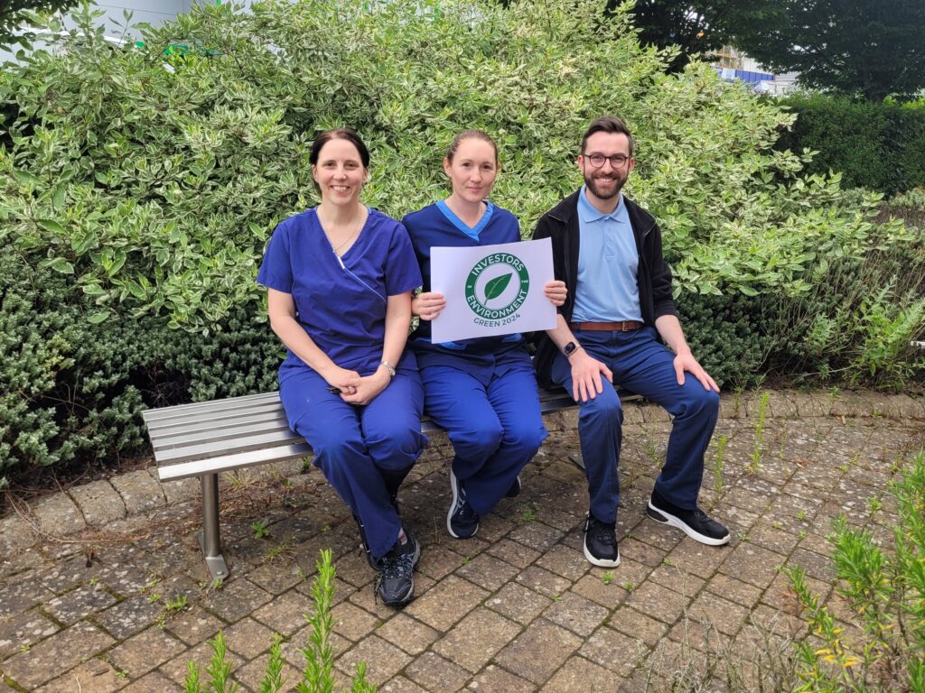 An image of three Willows associates sitting on a bench in a garden. From left to right is a white woman with dark hair wearing blue nursing scrubs, a white woman with res hair wearing blue nursing scrubs and holding a sign with the logo of investors in the environment printed on it, and a white man with short dark hair wearing dark blue trousers, a light blue polo shirt and black jacket. He is also wearing glasses. All three are looking at the camera and smiling.